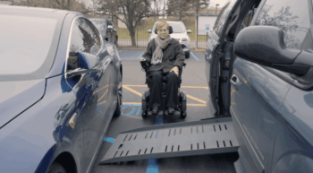 Liz is sitting in a power wheelchair in a parking lot sitting between her adapted van and a car that illegally parked in the striped access aisle blocking her from entering her van.