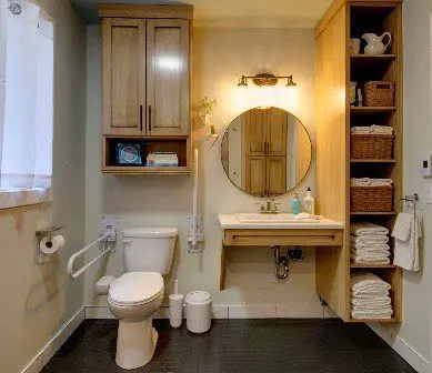 A picture of an accessible bathroom with a toilet located in a corner with two drop-down grab bars on each side, and enough room next to it so one could transfer from a wheelchair, and a sink with room for a wheelchair to roll under it. It also has storage for towels that goes low to the ground and can be reached by a wheelchair.