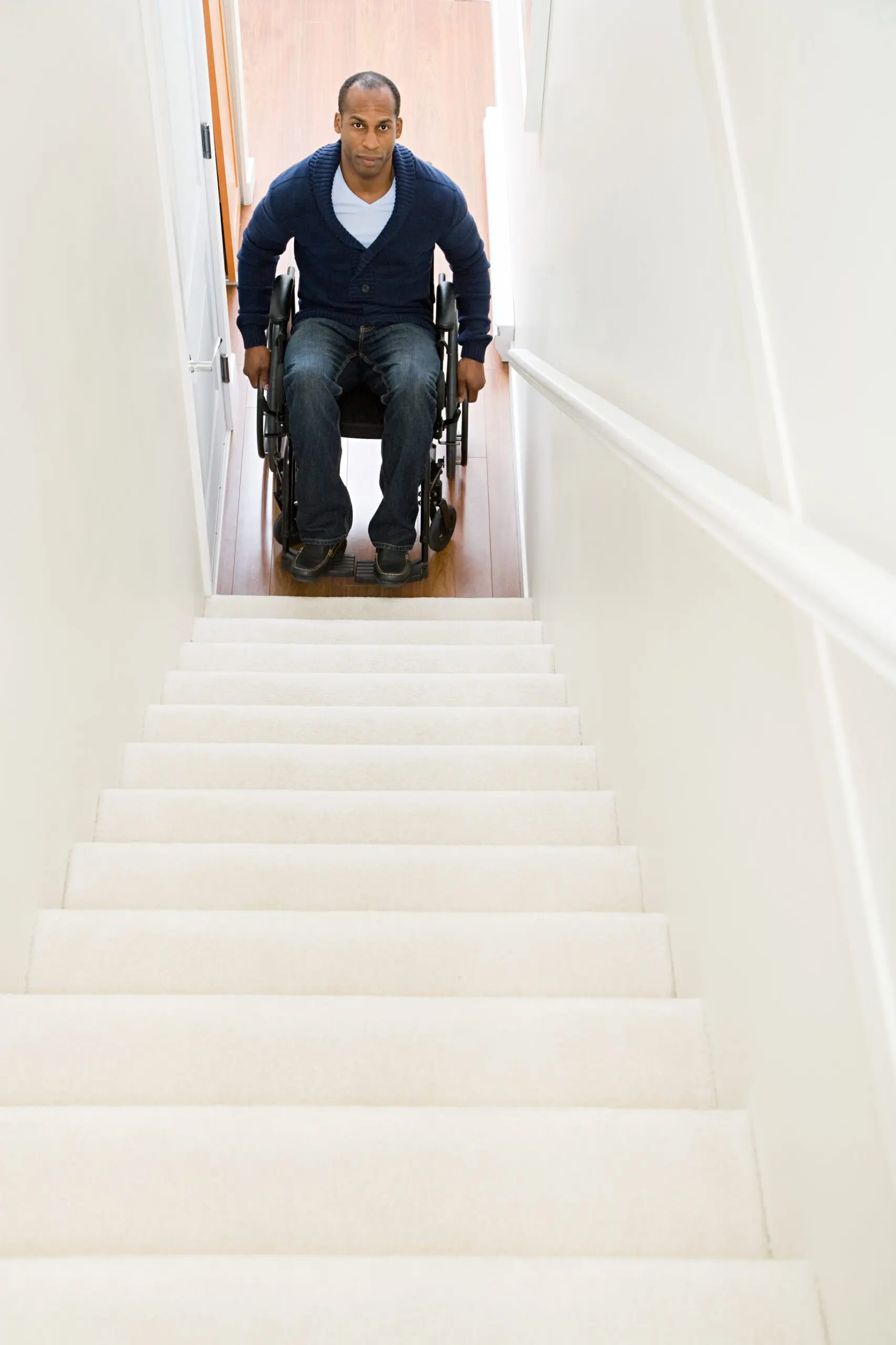 A Black man is sitting in a manual wheelchair at the bottom of a stairway looking up.