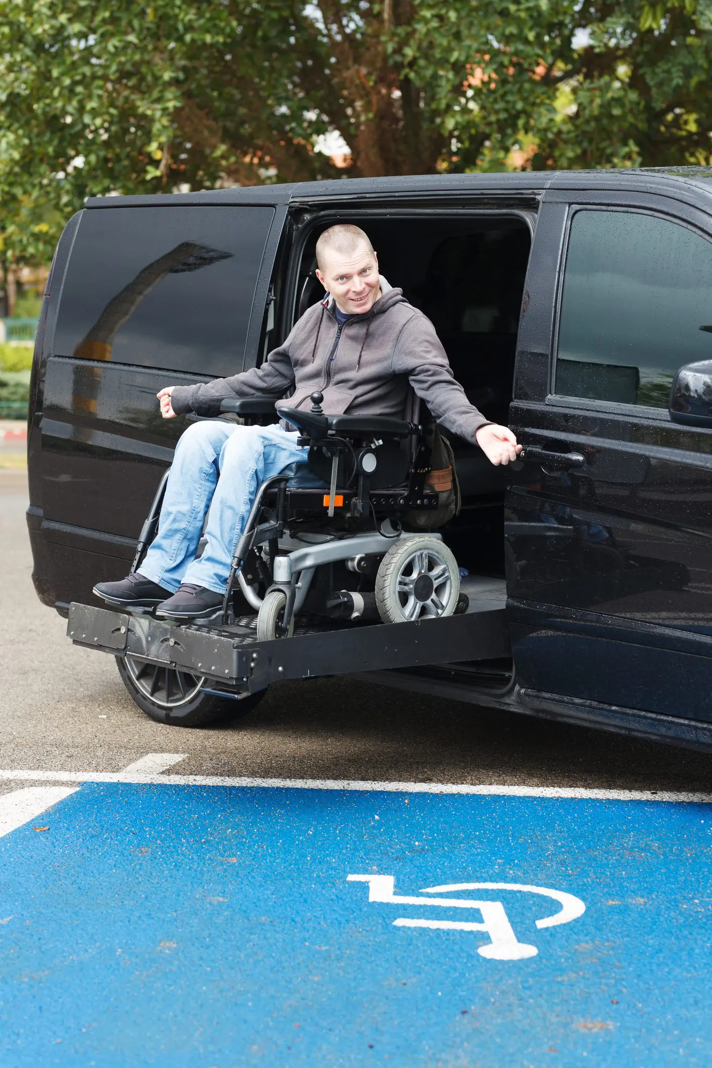 A white man in a power wheelchair is sitting on the lift of his adapted van.