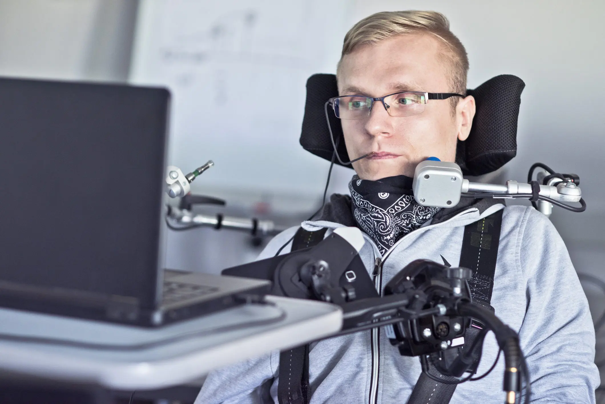 A White man sitting in a power wheelchair with using a mouth-directed mouse to operate a laptop computer
