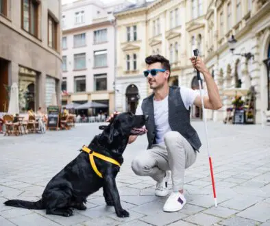 A blind White man is kneeling down to pet his black service animal in an outdoor plaza