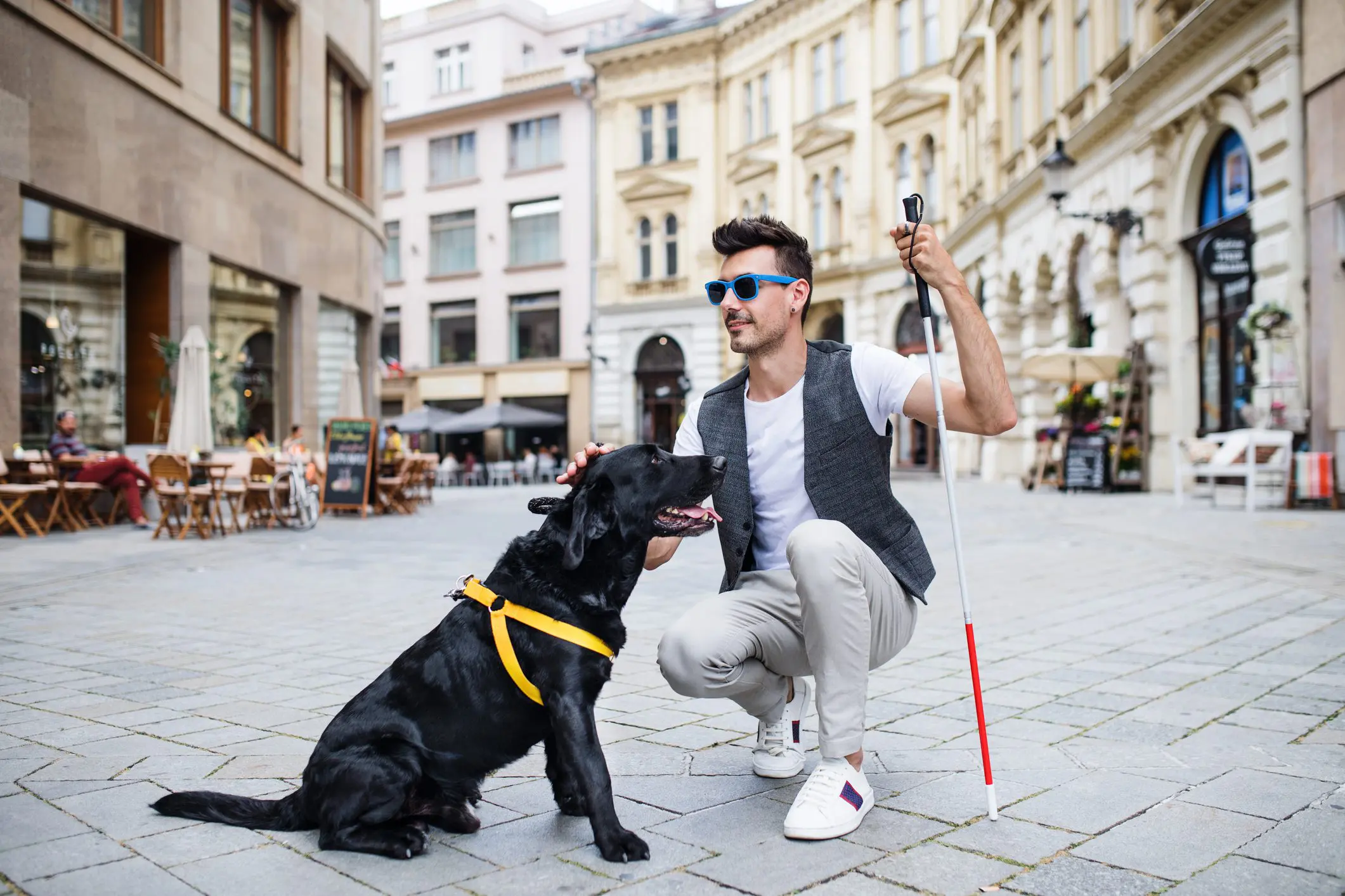 A blind White man is kneeling down to pet his black service animal in an outdoor plaza