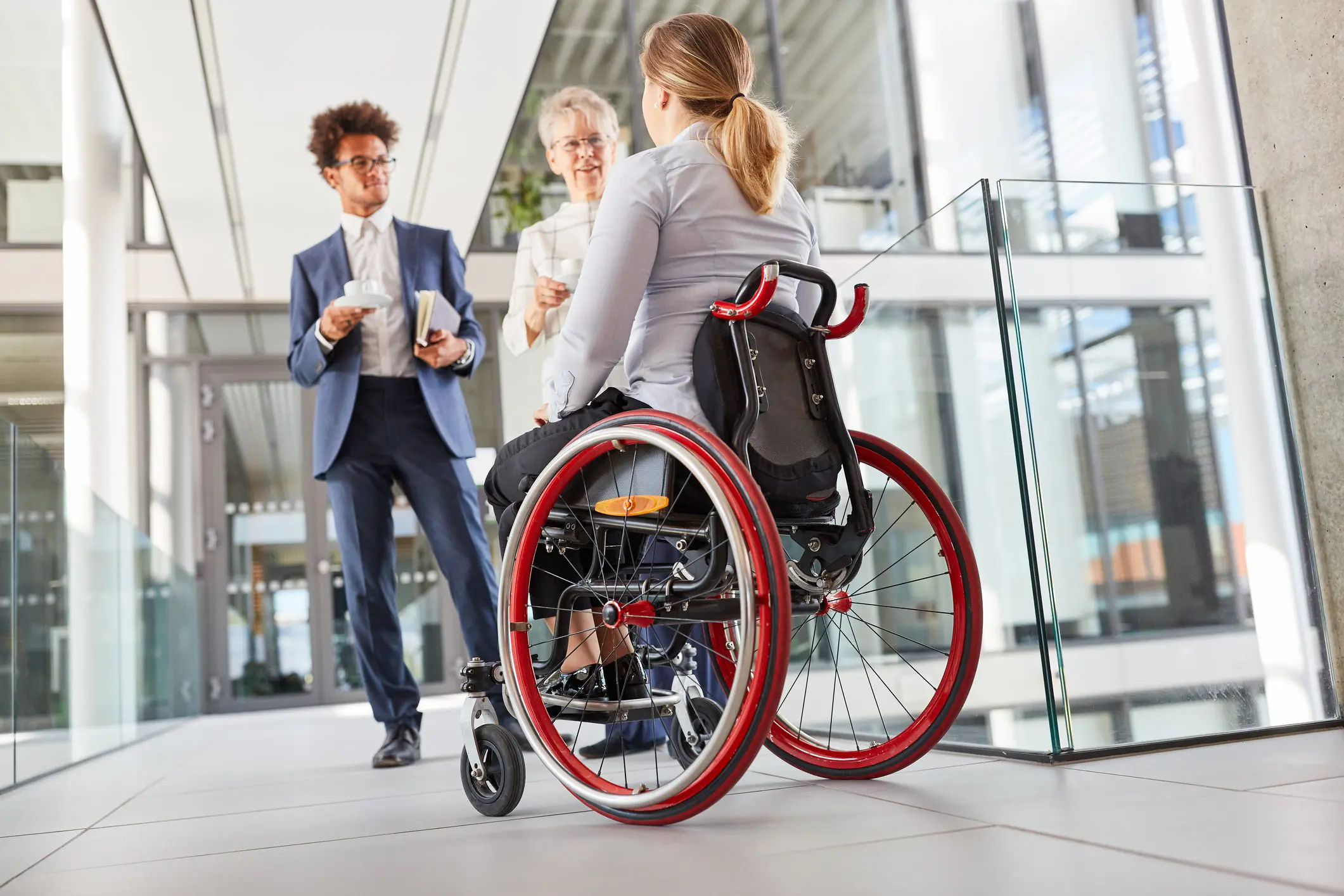 A white woman in a manual wheelchair with red rims talks to work colleagues who are a White woman and a Black man