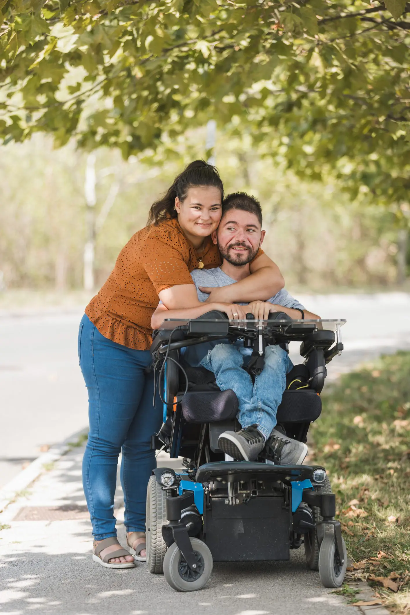 A fair-skinned woman is standing with her arms around a man sitting in a power wheelchair
