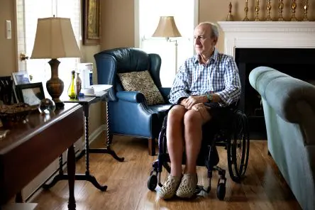 a White 70-year-old man with grey hair sitting in his living room in a manual wheelchair wearing a checked button-down shirt and shorts and looking out the window with a smile on his face.