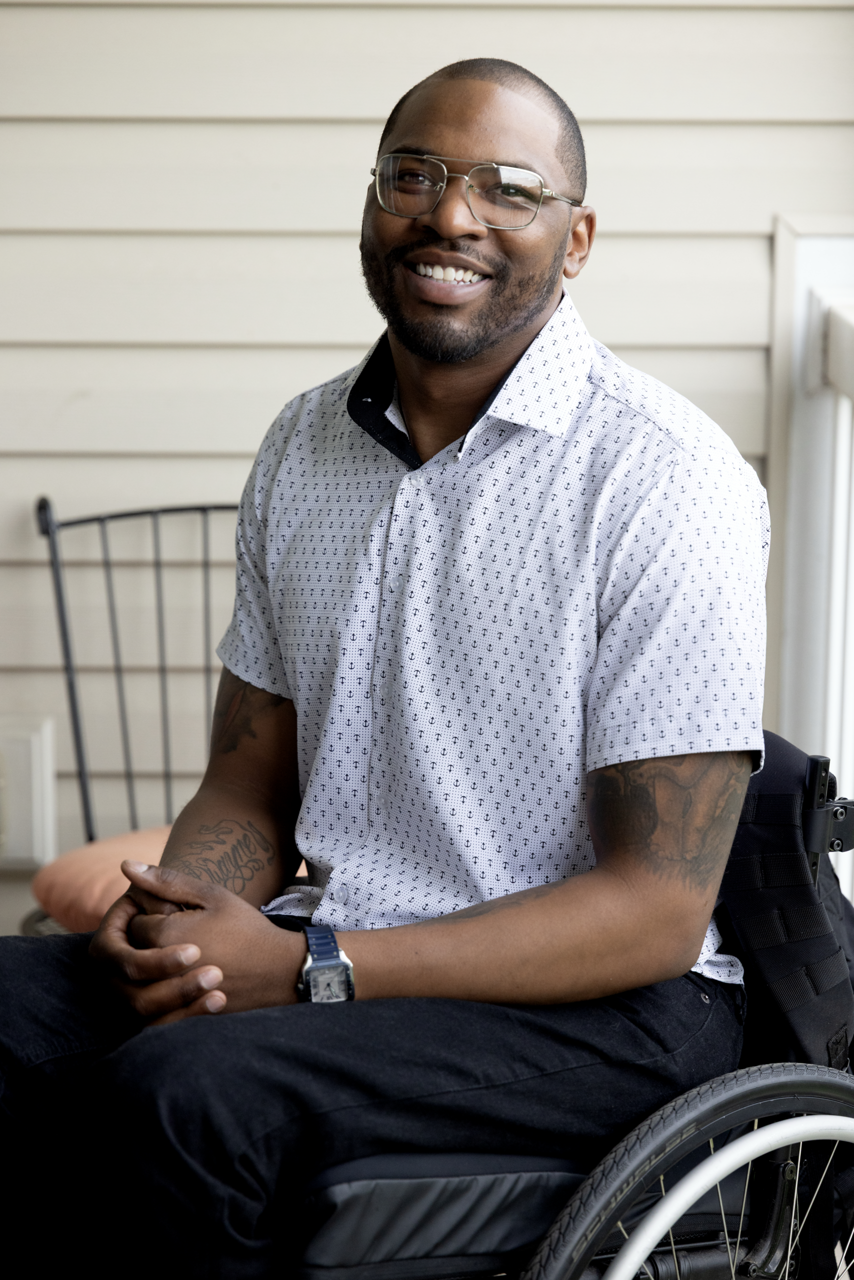 Terrell is a Black man sitting in a manual wheelchair on the porch of his apartment. He is wearing glasses, a white short-sleeved shirt and dark pants, and he's smiling at the camera.