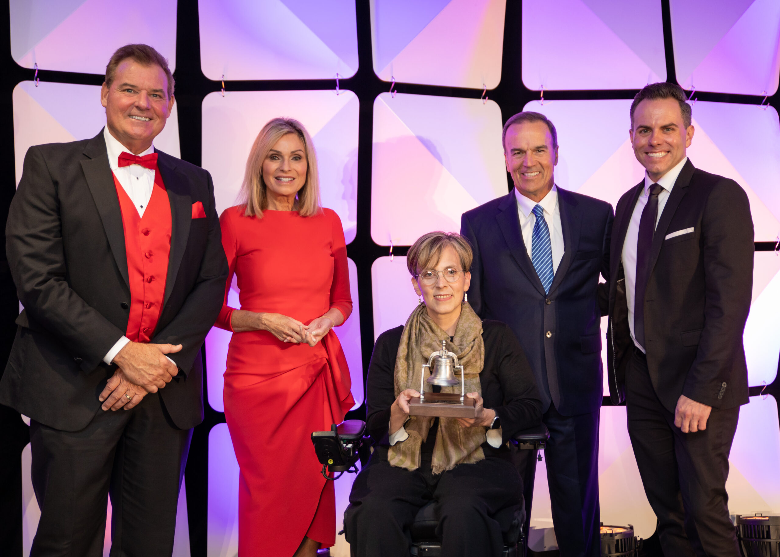 A smiling woman seated in wheelchair holding a Bell Award flanked by four local TV personalities, two on each side.