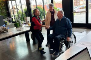 Person using a wheelchair shaking hands with someone standing, both smiling warmly.