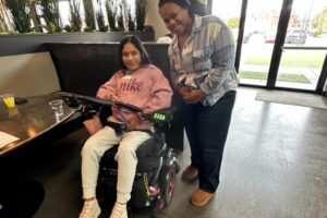 Young attendee using a power wheelchair and another individual smiling together next to a table.