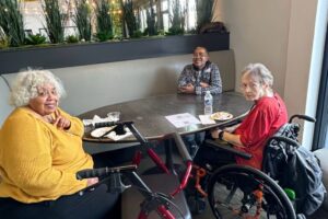 Three attendees, one with a mobility aid, chatting and sharing food at a table.