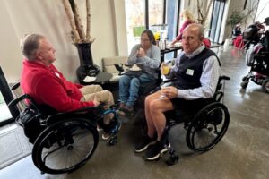 Two individuals using wheelchairs enjoying food and conversation in a bright venue.