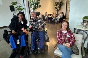 Two attendees using power wheelchairs and one seated in a chair smiling together.