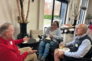 Three attendees, two using wheelchairs, talking and enjoying refreshments near windows.