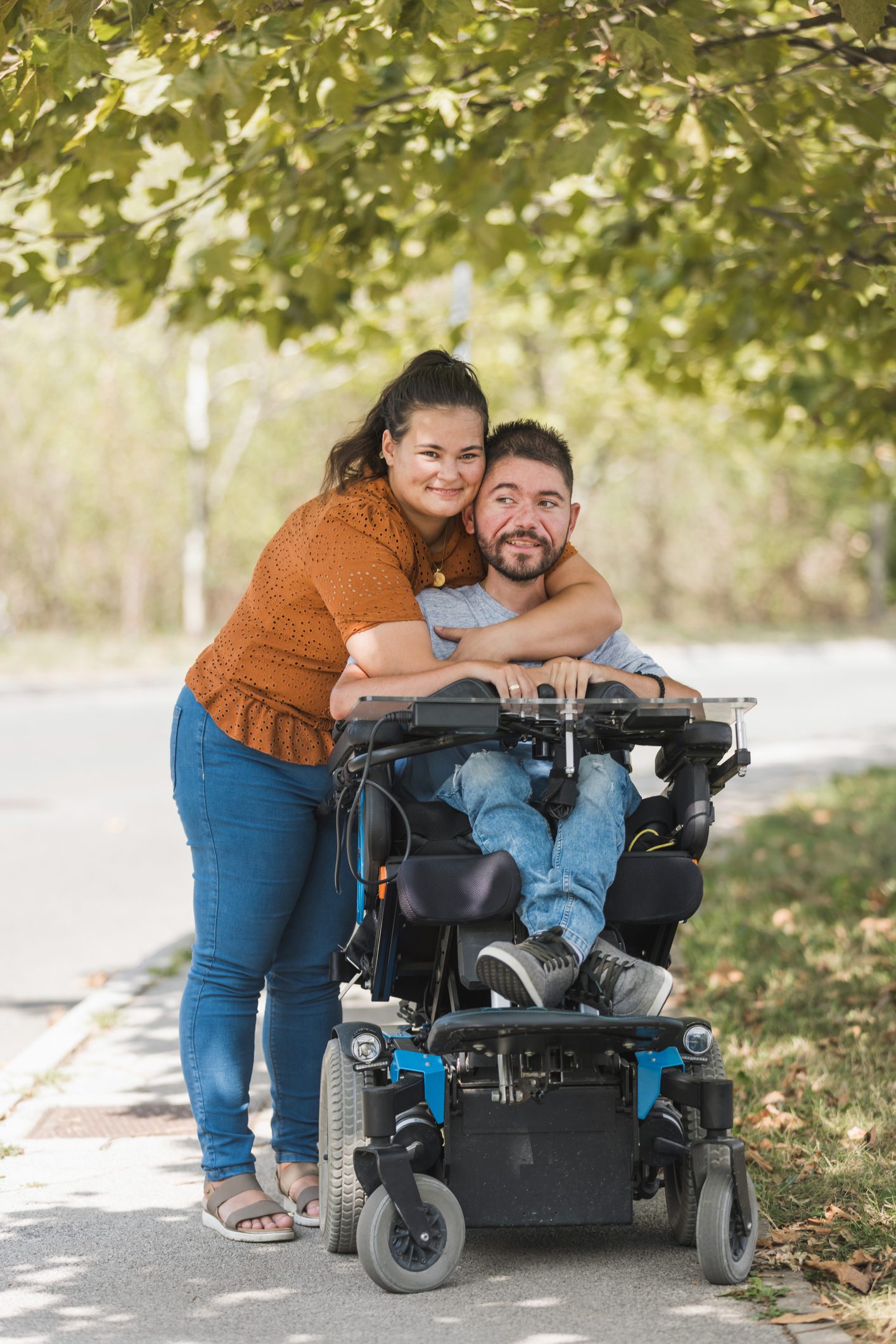 dreamstime_m_322410616(1) A fair-skinned woman is standing with her arms around a man sitting in a power wheelchair