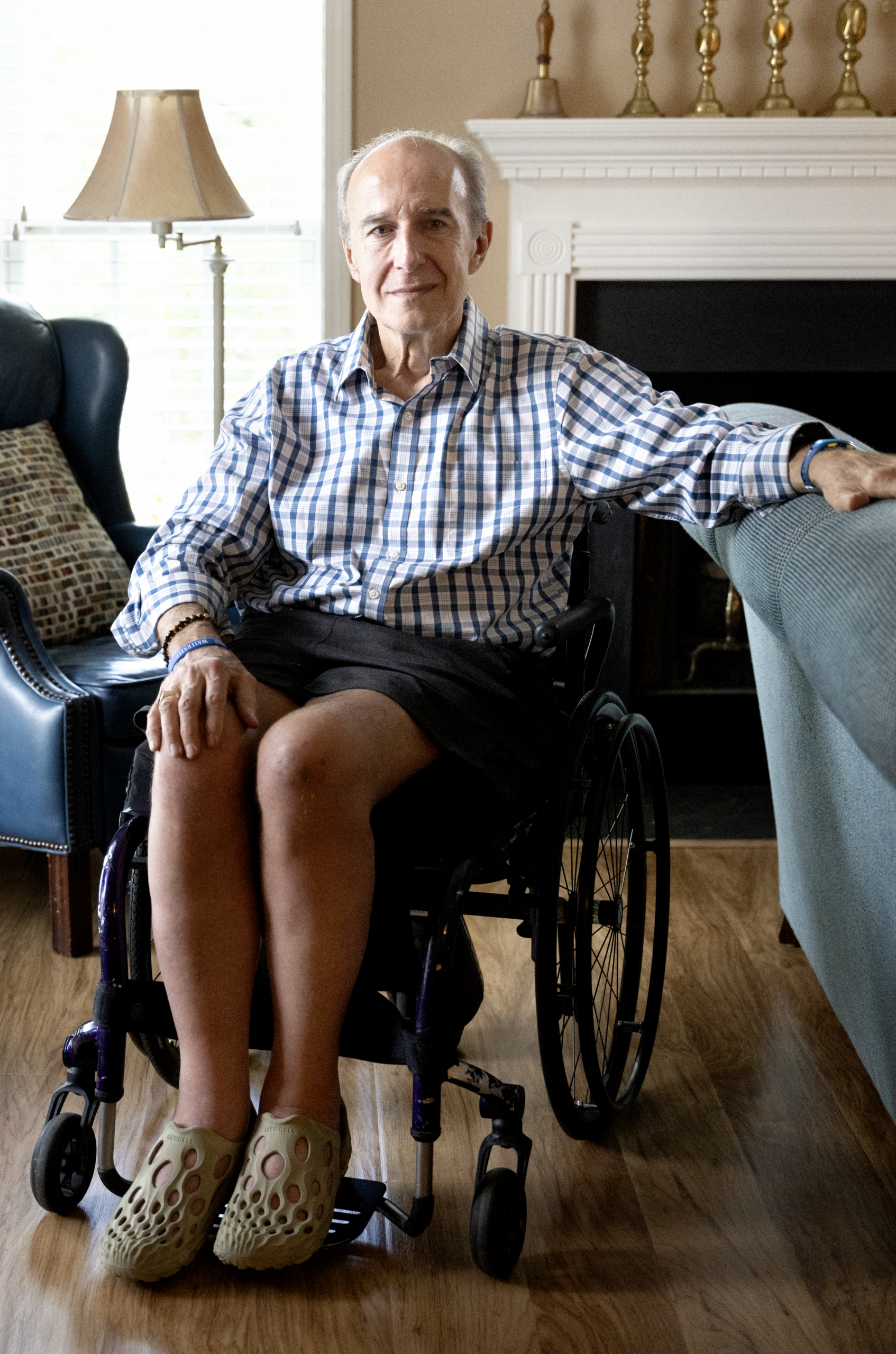 Garrison is a White 70-year-old man with grey hair sitting in his living room in a manual wheelchair wearing a checked button-down shirt and shorts, and looking straight at the camera with a serious expression.
