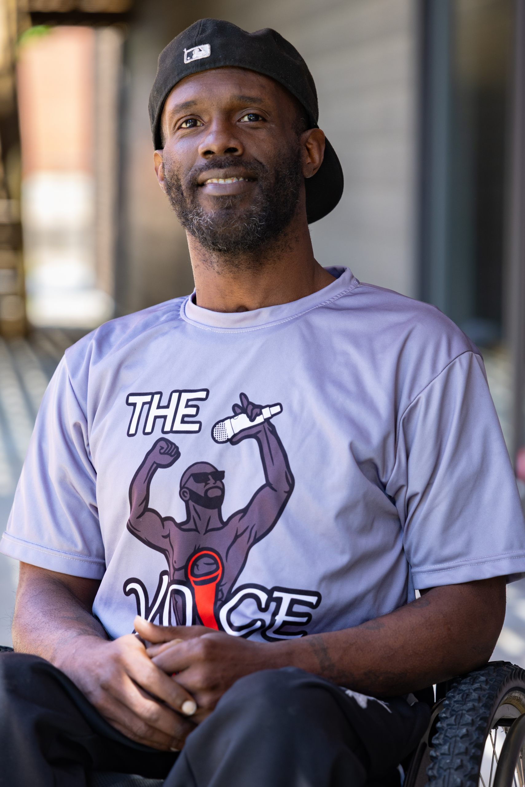 Kaelin is a Black man who is sitting in a manual wheelchair outside, wearing a light purple t-shirt smiling at the camera