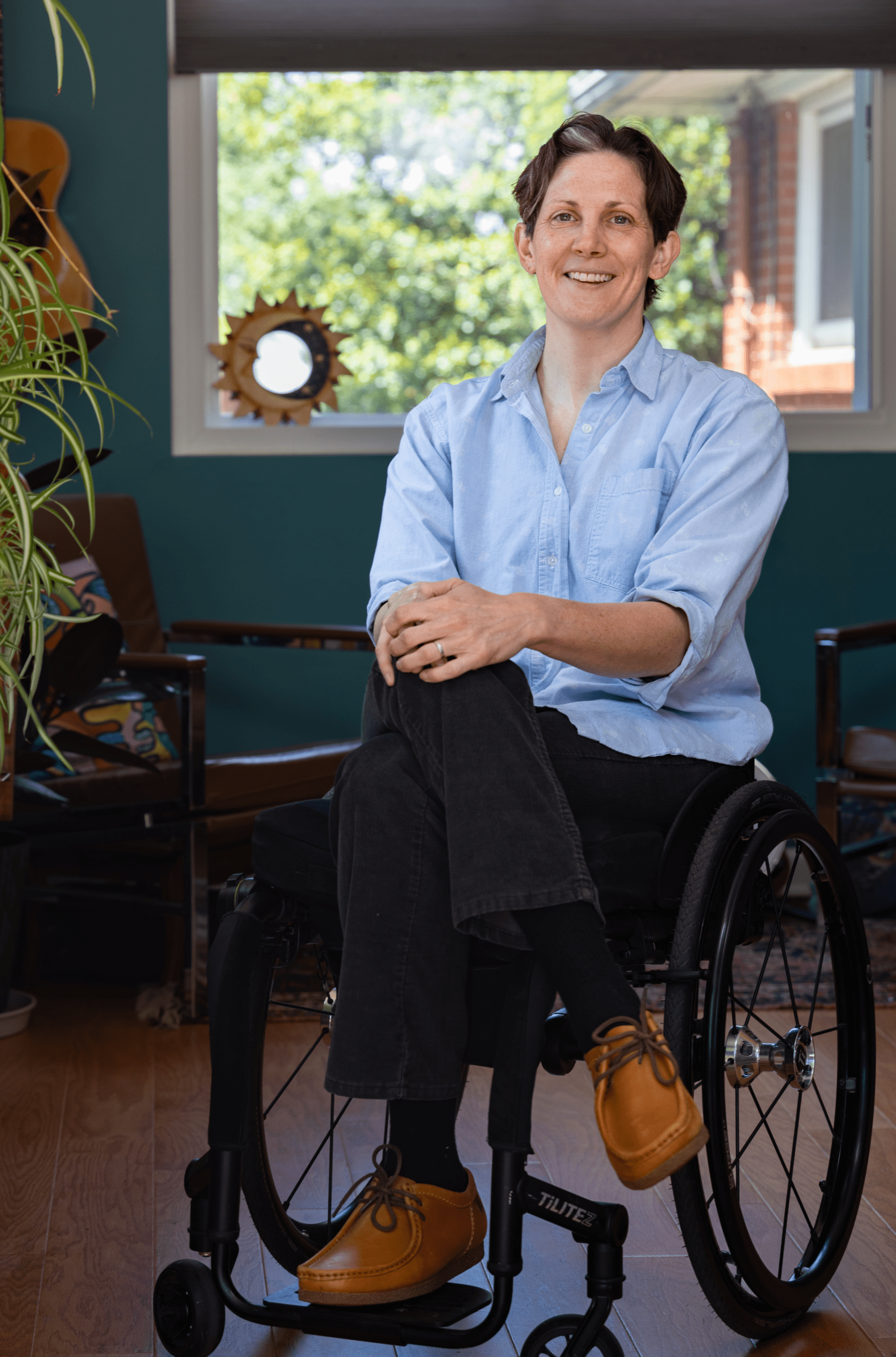 Rachel is a White woman sitting in her home in a manual wheelchair wearing a blue button-down shirt, and dark pants looking straight at the camera with a smile on her face.