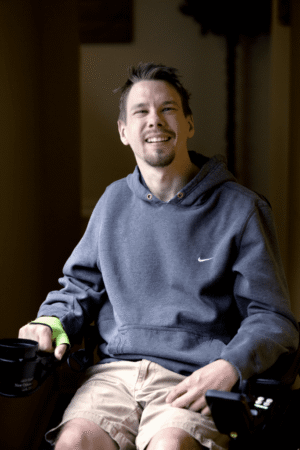 raham is a young White man sitting in a power wheelchair and wearing a dark grey hoodie, looking straight at the camera and smiling.